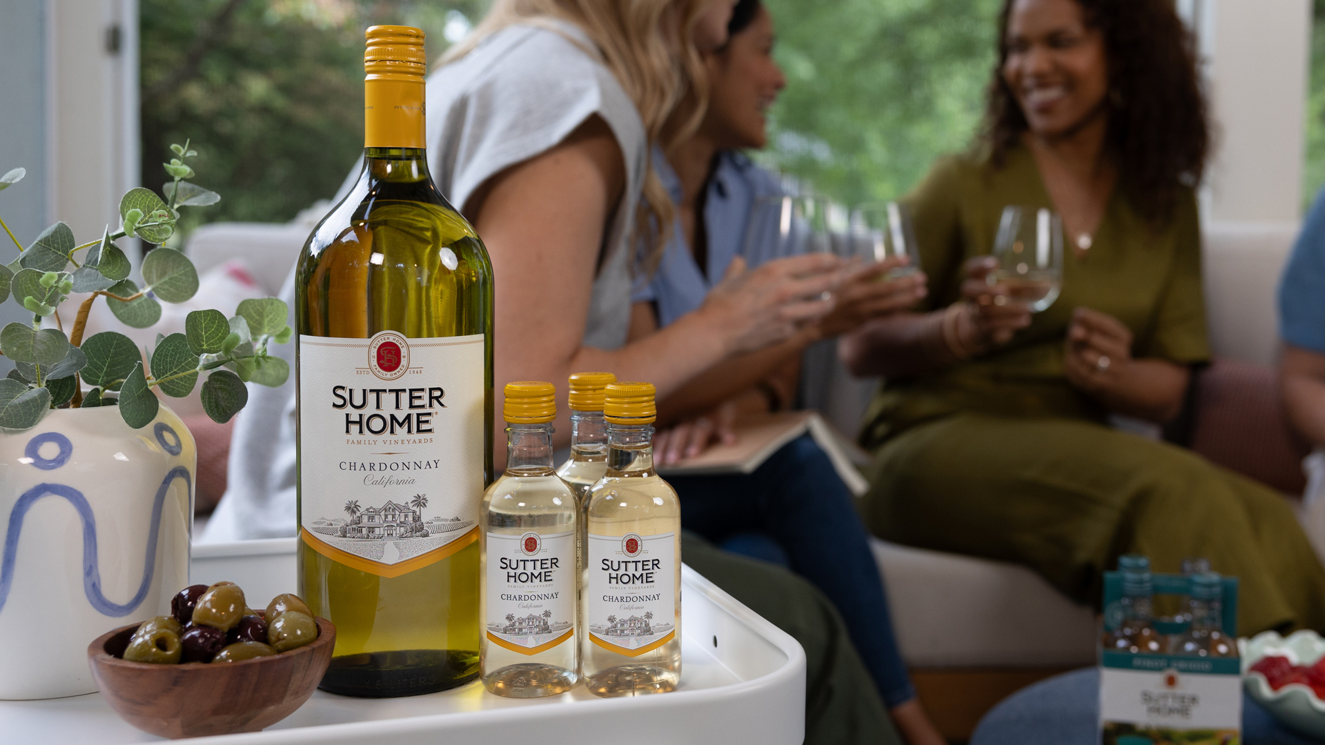 At home in the living room with Sutter Home Chardonnay large format bottle and mini bottles with a bowl of olives and a potted plant in the foreground, with three female friends in the background, sitting on a couch and holding wine glasses while smiling and chatting, in front of a big picture window.