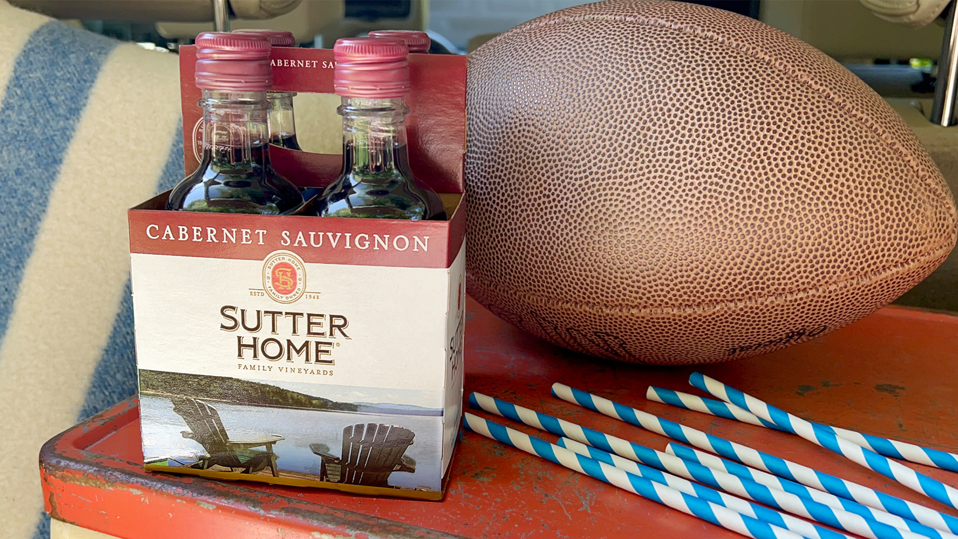 A table with a 4-pack of Sutter Home Cabernet Sauvignon mini bottles, a football, and a stadium blanket in the background - multiple items needed to take to the big game.