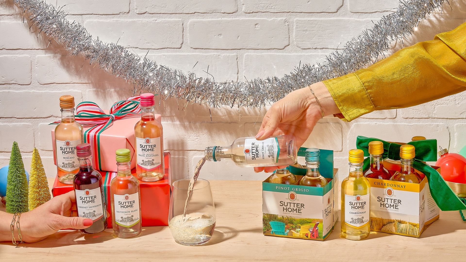 At home in the living room with Sutter Home Chardonnay large format bottle and mini bottles with a bowl of olives and a potted plant in the foreground, with three female friends in the background, sitting on a couch and holding wine glasses while smiling and chatting, in front of a big picture window.
